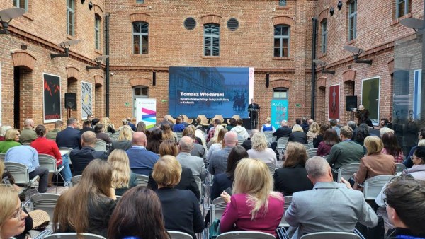 Szeroki kadr z widokiem na pełną widownię Małopolskiego Forum Ochrony Zabytków. W głębi na scenie przemawia Tomasz Włodarski, dyrektor MIK. W tle widać wielki ekran i zabytkowy ceglany dziedziniec Muzeum AK.
