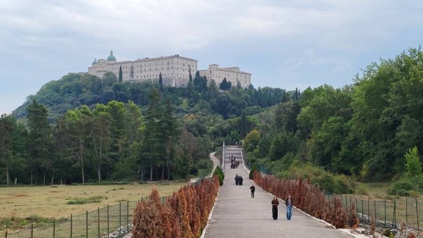 Długa, prosta droga prowadząca pod górę w stronę zalesionego wzgórza z zabytkowym klasztorem na Monte Cassino górującym nad okolicą. Po alei spacerują nieliczni przechodnie, a po bokach rosną szpalery tui i zielone drzewa.