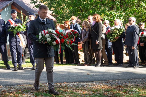 Uroczystości rocznicowe w Dąbrach. Na pierwszym planie mężczyzna w ciemnej kurtce i krawacie niesie wieniec z białych i czerwonych kwiatów z szarfą Muzeum AK. W tle widać czekających w szeregu uczestników, młodzież w mundurach i zieleń drzew.